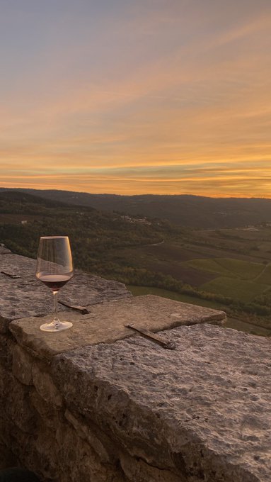 A glass of red wine sits on a stone wall overlooking rolling Tuscan-style hills and vineyards at golden hour sunset.