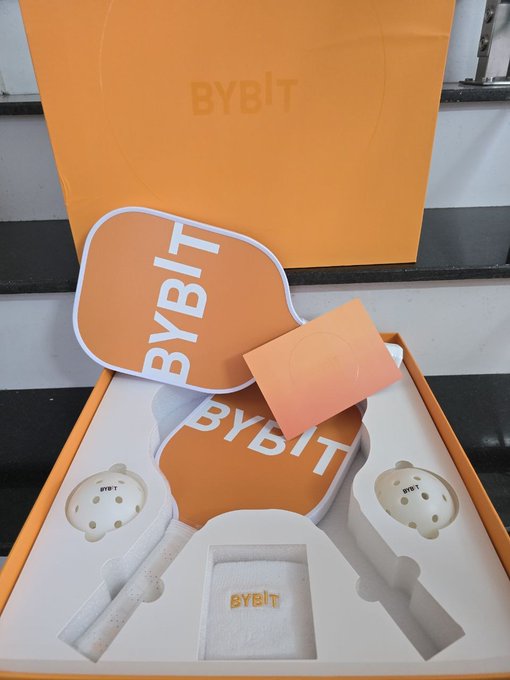 An orange Bybit-branded gift box is opened to reveal a Mooncake Festival set, featuring a paddle-shaped sign and two ping pong paddles with "BYBIT" printed on them, alongside a small orange packet. The image directly complements the post's message about receiving a thoughtful Mid-Autumn Festival gift from Bybit, showcasing the brand's signature orange color and attention to detail in its presentation.