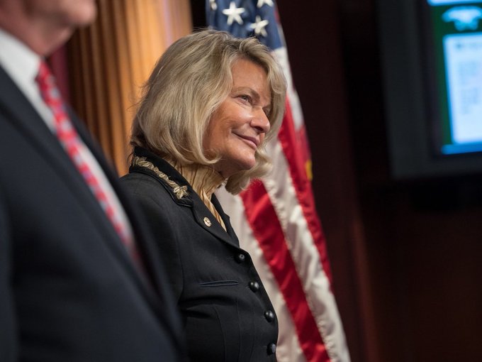 A woman with blonde hair, wearing a dark blazer and a lapel pin, smiles while standing in front of an American flag at what appears to be a formal event, with another person in a red tie partially visible in the foreground. The image corresponds to Senator Cynthia Lummis, who is featured in the post criticizing Democrats for allegedly stalling the CLARITY Act.