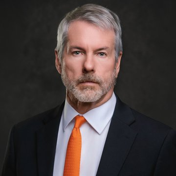 A distinguished man with gray hair and a beard, wearing a dark suit, white shirt, and orange tie, poses for a formal portrait against a dark background, appearing as a professional headshot likely accompanying the financial news about TD Cowen's reiterated buy rating on Strategy (MSTR).