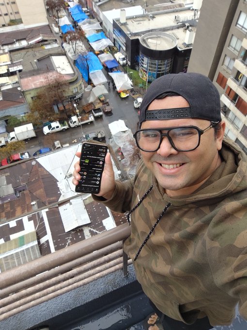 A man wearing a black cap, glasses, and camo hoodie takes a selfie on a high balcony while holding up a smartphone showing a chat app, with a city street market visible far below.