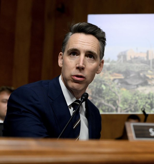 A man in a dark suit and striped tie speaks at a Senate hearing, seated in front of a microphone with a screen showing an image of the Great Wall of China behind him.