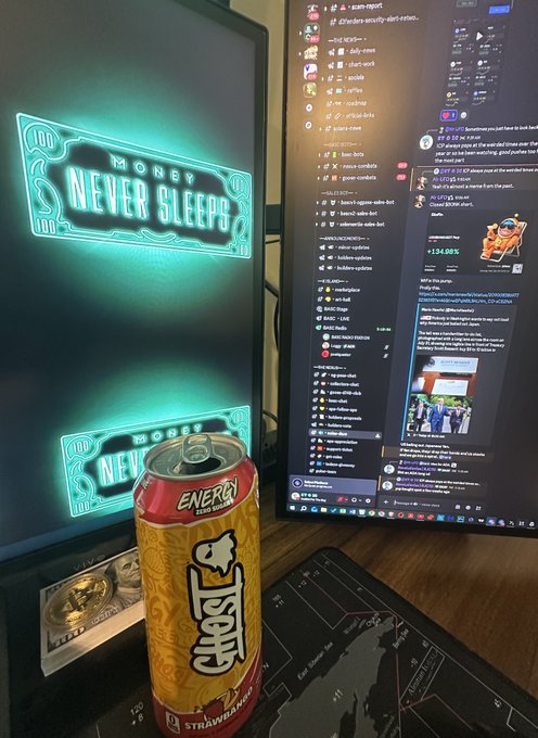 A desk setup featuring a can of 'Ghost Energy' Strawberry banana flavor, with neon signs reading 'MONEY NEVER SLEEPS' and a monitor displaying a crypto trading Discord chat.
