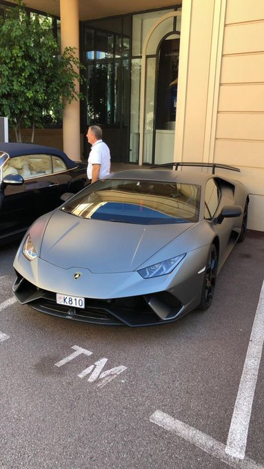 A matte gray Lamborghini Huracán with license plate K810 is parked in front of an upscale building, with a man in a white shirt standing nearby.