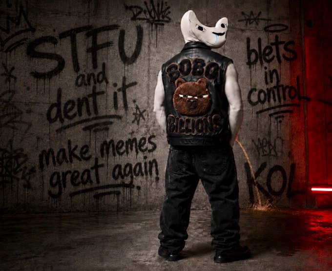 A person wearing a white mask and a leather vest with 'BOBO BILLIONS' and a bear patch, standing in front of a graffiti wall reading 'STFU and dent it. make memes great again.'