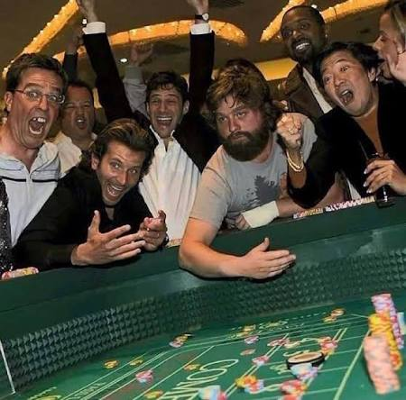 A group of excited men, including Bradley Cooper, are gathered around a craps table at a casino, cheering and throwing their arms up in celebration.