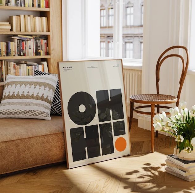 A framed art print leaning against a wall in a cozy room, featuring a minimalist geometric design with black shapes, a white circle, and an orange dot, labeled 'MOCKBA' and 'blue perspective'.