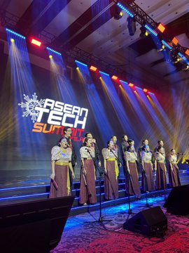 Seven performers in white and red traditional attire stand on stage at the 'ASEAN TECH SUMMIT' event with dramatic stage lighting.