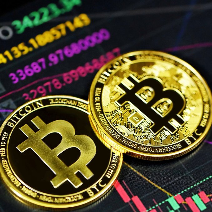 Two golden Bitcoin tokens rest atop a digital display showing Bitcoin's price near $34,221 and candlestick chart patterns. The image visually reinforces the article's point about BTC's current market positioning relative to traditional assets, illustrating the cryptocurrency that K33's analyst says still has potential upside compared to gold and equities.