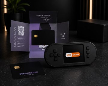 The image shows an open seedkeeper wallet with a black screen and buttons, sitting on top of a table next to two small cards - one purple and one orange. The text on these cards reads "Seed Keeper" and "Seed Keeper Seed Keeper". The wallet is placed in front of a potted plant, which adds a touch of greenery to the scene. The image also contains some text that says "It's finally here. After a long journey we want thank the open source community for progressing to this point."
