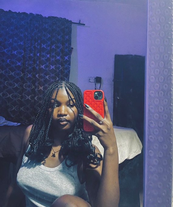 A young woman with long black braided hair takes a mirror selfie in a dimly lit purple room using a red phone case.