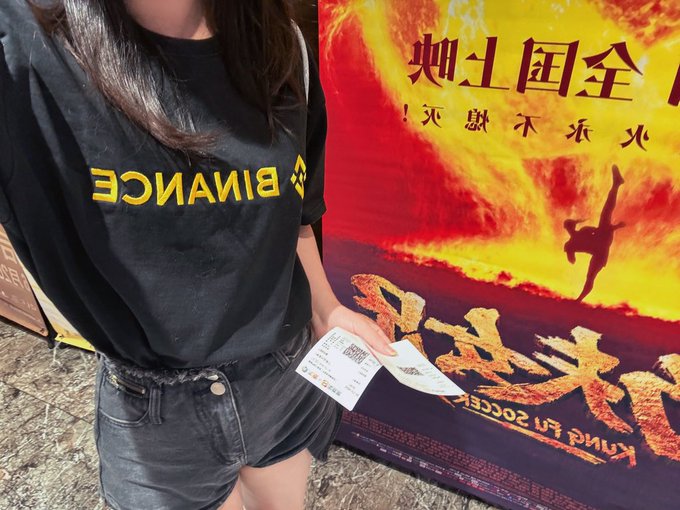 A woman wearing a black 'BINANCE' t-shirt holds two tickets in front of a movie poster with Chinese text and a release date of 'AUGUST 2, 2024' visible.