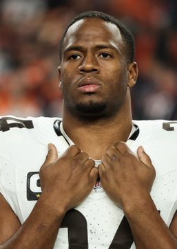 Nick Chubb, wearing his Cleveland Browns #24 white jersey, stands with his hands over his heart in a solemn pregame pose. The image captures the longtime Browns running back during his playing days, fitting as he returns to Cleveland to retire as a member of the organization where he spent the prime of his career.