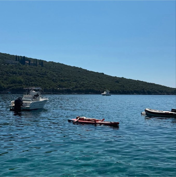 A serene coastal scene with clear blue water where a person is relaxing on a floating pink and black inflatable pad, with a small motorboat anchored nearby and another boat in the distance against a backdrop of green forested hills.