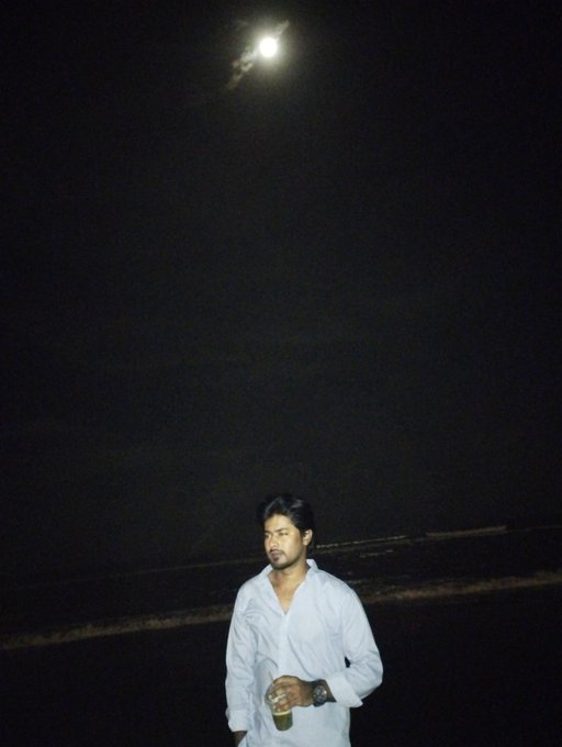 A nighttime photograph of a man in a white shirt holding a cup, standing against a dark sky with the moon visible above him.
