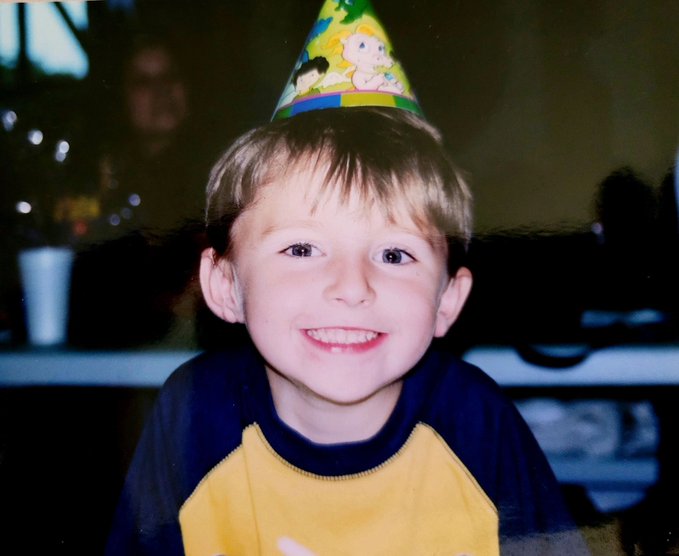 A photograph of a smiling young boy wearing a yellow and blue shirt and a colorful, cartoon-decorated birthday party hat.