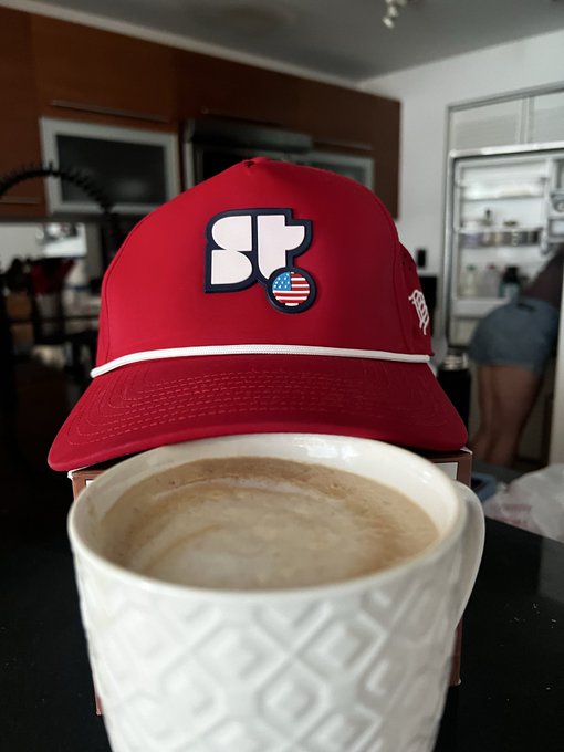 Red baseball cap with 'STR' logo and American flag patch placed behind a white textured mug filled with coffee, set in what appears to be a cafe or kitchen setting.