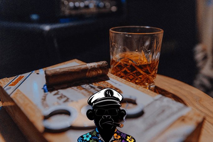 A glass of whiskey with ice sits on a wooden surface alongside a cigar, a cigar cutter, and a decorative box, with a cartoon monkey wearing a captain's hat and Hawaiian shirt edited into the foreground.