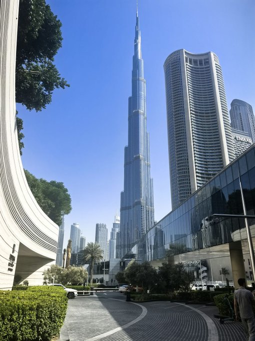 A daytime photograph of the Burj Khalifa skyscraper in Dubai, towering above surrounding buildings including the 'EMAAR' Address tower against a clear blue sky.