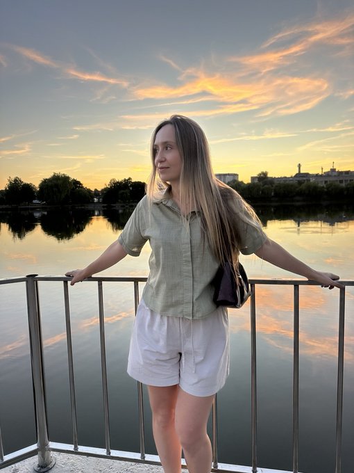 A young woman with long blonde hair, wearing a light green shirt and white shorts, standing on a pier at sunset, looking out over a body of water.