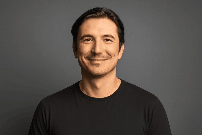 A portrait of a smiling middle-aged man with dark hair wearing a plain black t-shirt against a neutral grey background.