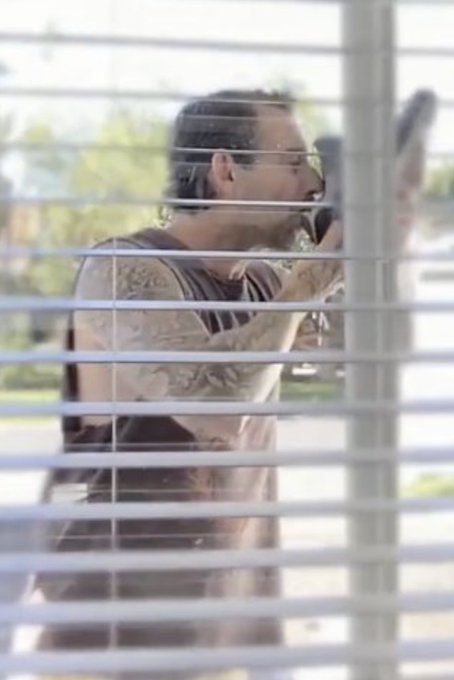 A bearded man with glasses and tattoos, wearing a sleeveless shirt, is photographed through horizontal window blinds while talking on a cell phone.