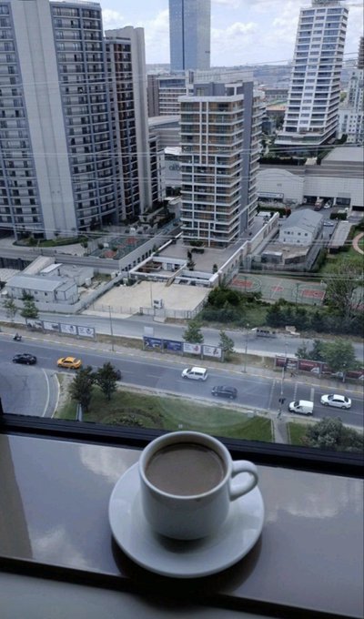 A view from a high-rise window showing a small espresso cup on a saucer in the foreground, overlooking an urban cityscape with modern apartment buildings, a road with cars, and greenery under a cloudy sky.