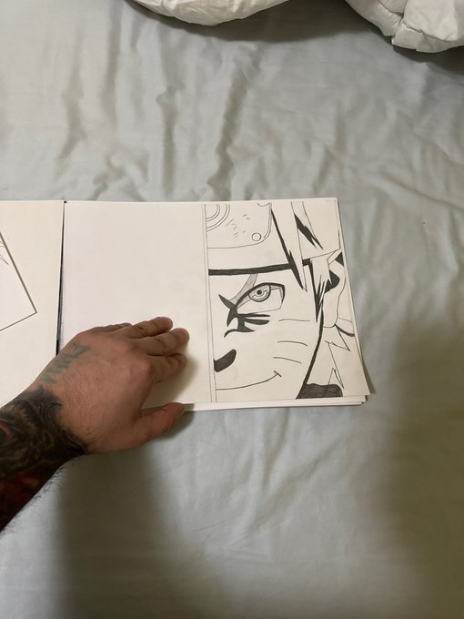 A tattooed hand rests on a manga sketchbook displaying a black-and-white drawing of the character Naruto with whisker marks on his face, lying on a white bedsheet.
