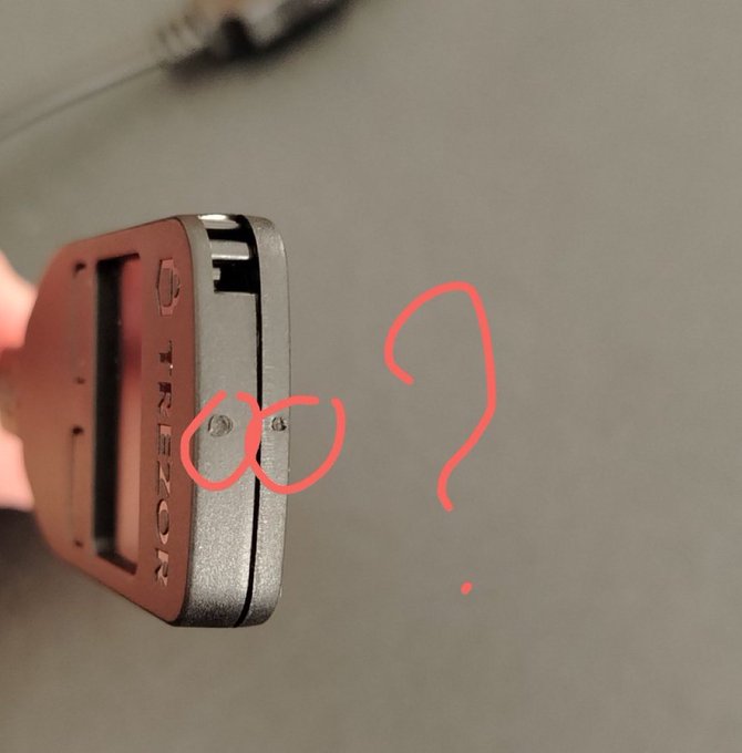 A hand holds a Trezor hardware wallet sideways, with red marker circles highlighting what appear to be small suspicious holes or modifications on the device's side, alongside a hand-drawn question mark. The image visually supports warnings about tampered Trezor devices in circulation, drawing attention to potential physical tampering that could compromise cryptocurrency security.