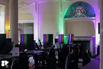 The image shows a venue being prepared for an event, with rows of computer monitors set up on desks and colorful purple, green, and blue stage lighting illuminating the room, featuring an ornate crest on the wall above. The setup aligns with the post about a hackathon or launch event hosted by @cstldao with @EasyA_Kickstart, ready for attendees to arrive as doors open at 9am.