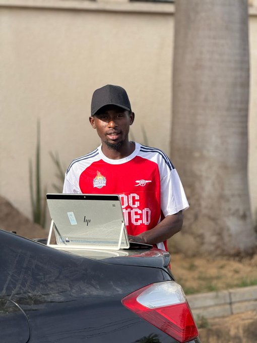 A man wearing a black cap and a red-and-white "SABON KARATU RISK MANAGEMENT" jersey stands behind an HP laptop placed on the trunk of a black car, appearing to explain the trading risk management system referenced in the post.