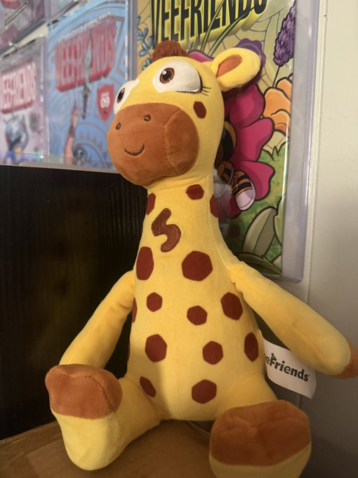 Plush toy of the character Geoffrey the Giraffe from Toys 'R' Friends, sitting in front of what appears to be Be@rbrick packaging.
