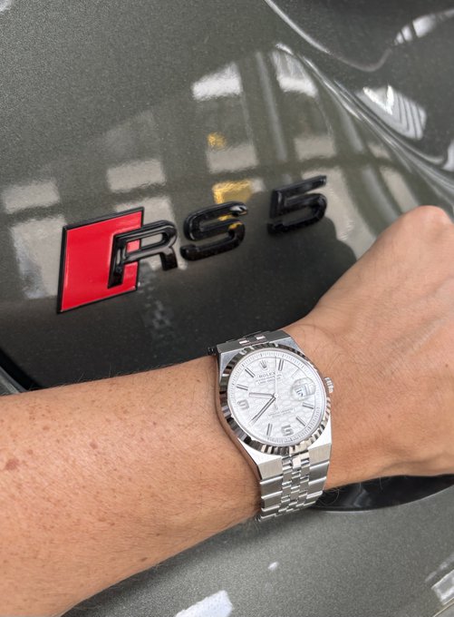 A close-up of a wrist wearing a silver Rolex Datejust watch next to a car with an 'RS 5' badge on its body.