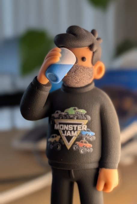 A 3D rendered figurine of a bearded man wearing a Monster Jam sweater drinking from a cup in an indoor setting.