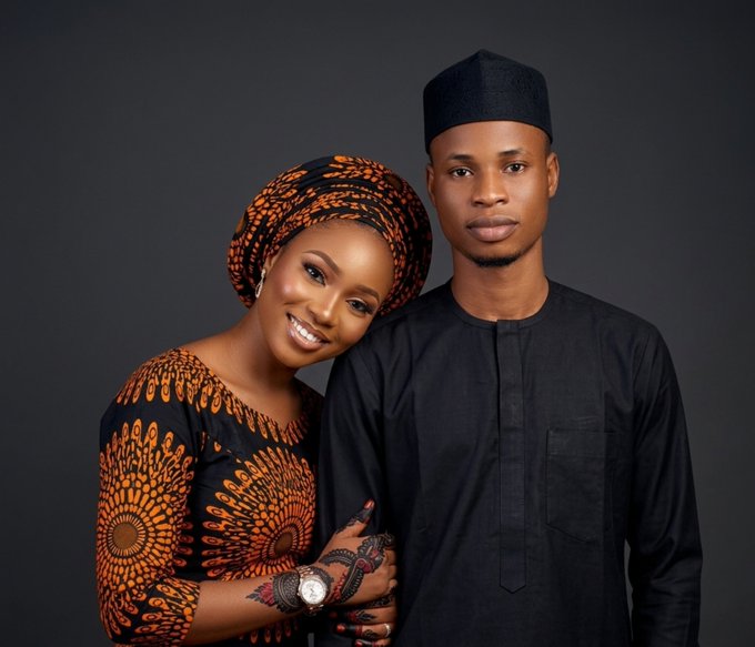 A studio portrait of a smiling couple in traditional Nigerian attire; the woman wears an orange and black patterned dress and headwrap, while the man wears a black outfit and cap.