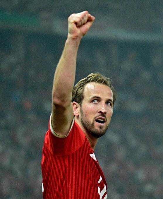 A photograph of soccer player Harry Kane in a red Bayern Munich jersey celebrating with a raised fist.