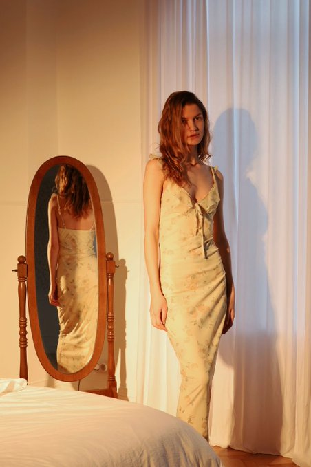 A woman in a floral silk slip dress stands beside an oval mirror that reflects her image, with white curtains and warm lighting in the background.