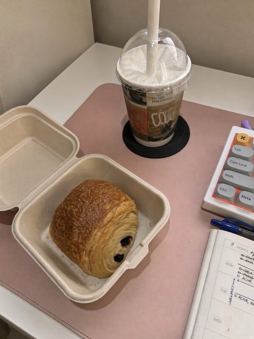 A pastry sits in an open compostable takeout container on a desk beside an iced drink, planner, pen, and a small educational device; the cup reads “NO ONE FIGHTS ALONE.”