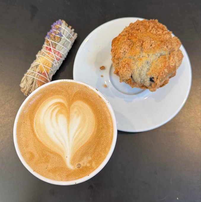 An overhead view of a coffee with heart-shaped latte art next to a large blueberry scone on a white plate, with a sage smudge stick resting on the dark table surface.