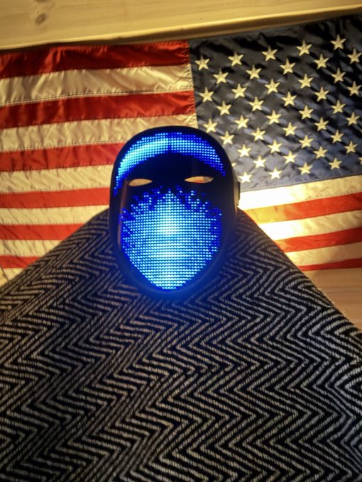 A blue LED-lit mask sits atop a black-and-white patterned surface with a U.S. American flag draped in the background, with no visible text or numbers. The post's boast about sharing "alpha" and profitable crypto picks like $KINS contrasts with the anonymous, unidentifiable face in the image—suggesting the poster's hype comes from a faceless source.