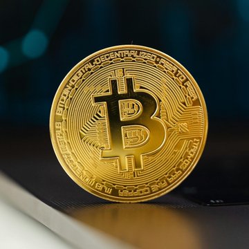 The image features a single gold Bitcoins worth $75,000 displayed on top of a table. The surrounding area seems to be a computer desk or a wooden table with a laptop on its surface. A person can be seen sitting at the desk, but their presence is not relevant to the context of Bitcoin's value in the market depicted in the image. 
The text "LATEST: Bitcoin fell below $75,000 on Wednesday as oil slid on US-Iran peace hopes, with BTC weakening even as US stocks hovered near record highs." explains that the price of Bitcoins dropped below $75,000 on a specific day. The reason for this drop might be attributed to changes in the oil market or other factors impacting the value of Bitcoin in the world economy.