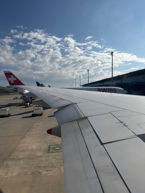 A view from an airplane window shows the wing of a SWISS aircraft and other planes with the Swiss cross logo on their tails, parked at an airport tarmac under a partly cloudy sky.