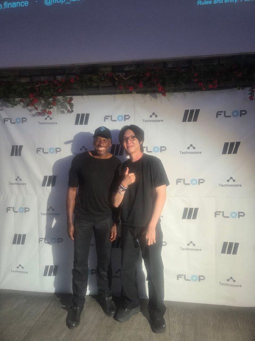 Two men pose together on a step-and-repeat backdrop for 'FLOP' and 'Technocore' events; the man on the left wears a blue cap and black t-shirt, while the younger man on the right wears glasses and a black t-shirt.