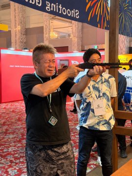 An older man with glasses, identified as Mr. Horie, playfully aims a toy rocket launcher at the camera while wearing a lanyard at the bitFlyer booth, with the booth's "Shooting" sign visible in the background. His enthusiastic pose at the cryptocurrency company's booth reflects the post's message about his enjoyable visit to explore their updated app and festival offerings.