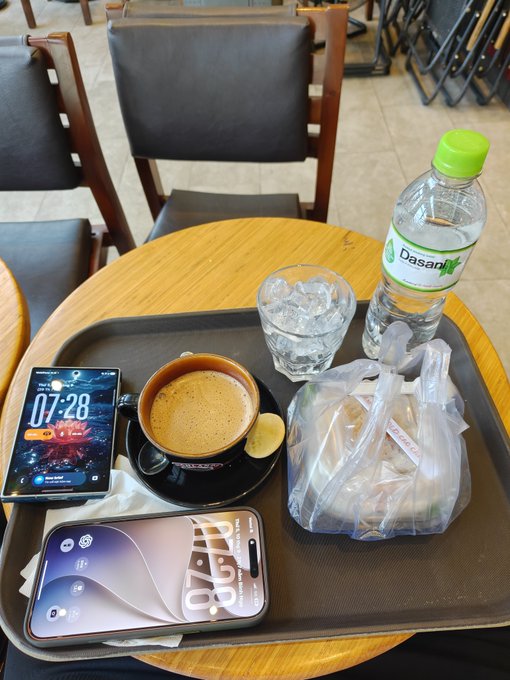 A tray on a wooden table at a cafe holds a coffee cup, a glass of ice water, a bottled water labeled 'Dasani', two smartphones (one showing '07:28'), and a plastic bag containing food.