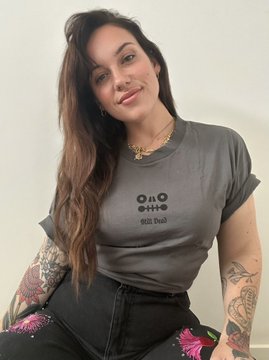 A woman with long brown hair and tattoos, wearing a gray "OO With Duck" t-shirt and black pants with pink floral embroidery.