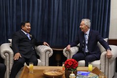 Two men in business suits sit in white armchairs in a formal meeting room, with glasses of tea and a red flower arrangement on a table in the foreground.