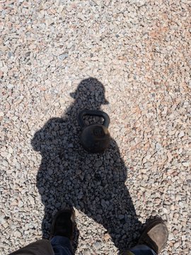 The shadow of a person wearing a baseball cap and carrying a backpack is cast on an asphalt surface, with the camera they are holding visible as a dark shape in the shadow's hands.
