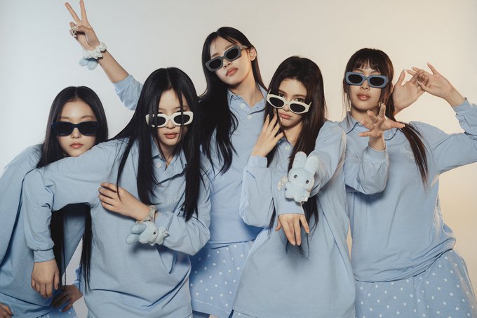 Five young women pose playfully in matching light blue outfits with sunglasses, one holding a small blue plush toy.