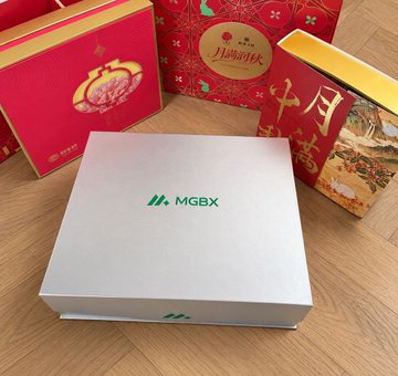 A silver metallic box branded with the green "MGBX" logo sits on a wooden surface, surrounded by several red and gold decorative gift boxes, likely Chinese mooncake packaging.